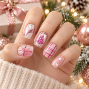 One hand wearing pink and white short square nails with a framed Christmas tree, ribbon bow, checks, hearts and a shimmering wave, beside pink Christmas decorations.