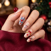 Hand wearing nude pink nails with raised cherries, a snowman and a wreath, beside a burgundy sleeve and blurred festive lights.