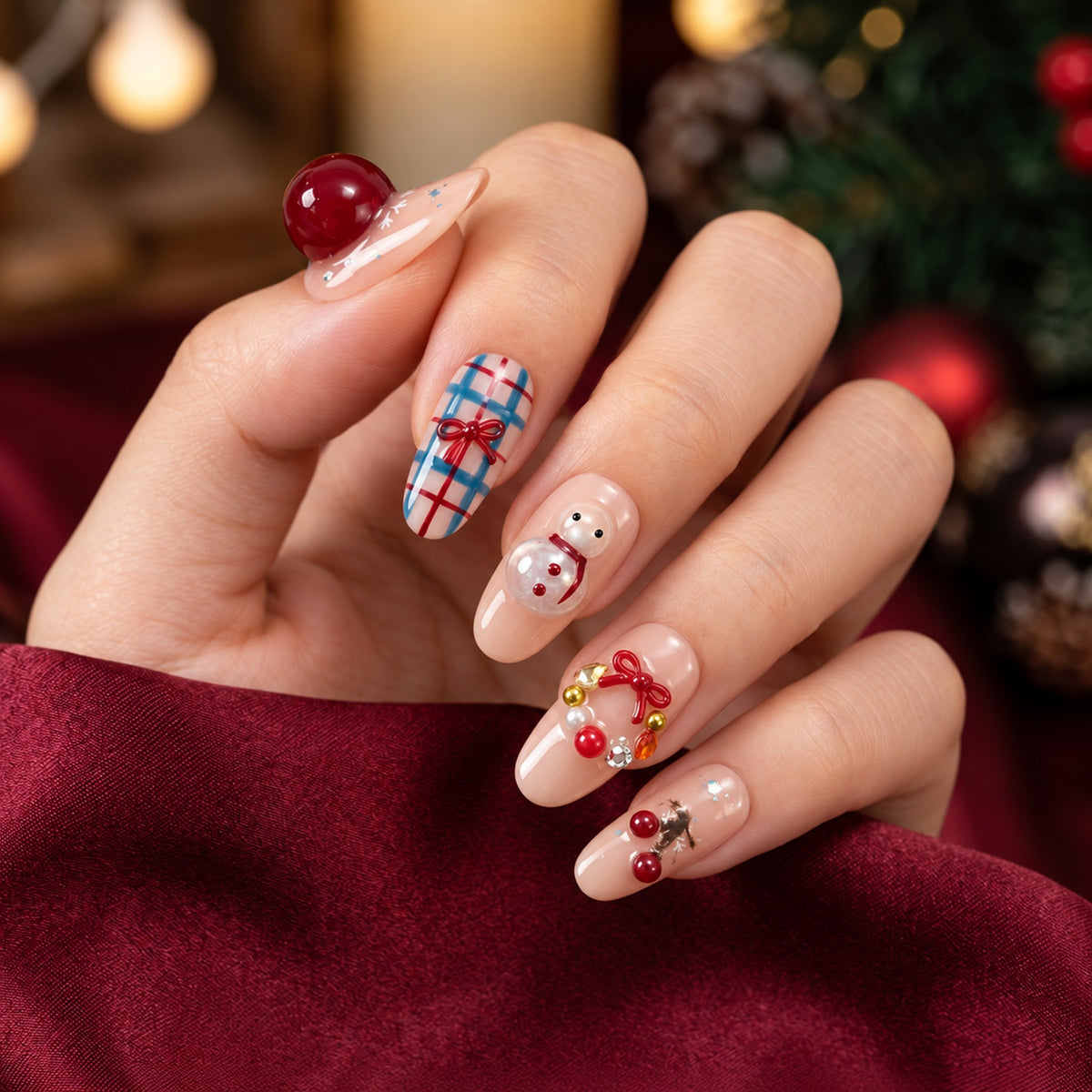 Hand wearing nude pink nails with raised cherries, a snowman and a wreath, beside a burgundy sleeve and blurred festive lights.